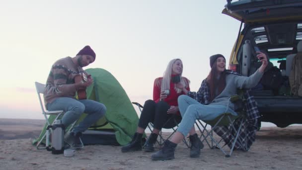 Jeune homme joue de la guitare et deux femmes s'assoient sur des chaises de camping-car, boit un thé chaud et prend selfie — Video