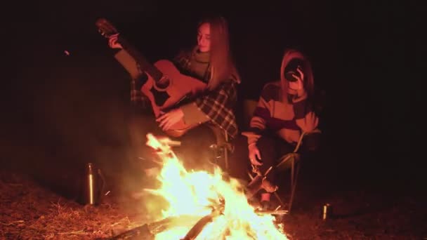 Дві молоді жінки з багаттям у лісі, вони грають на гітарі та співають пісні — стокове відео