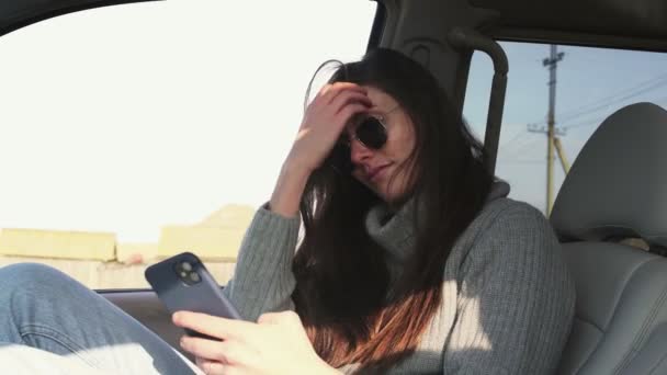 Jeune femme avec des lunettes de soleil est assis en voiture et prend selfie le matin d'été — Video
