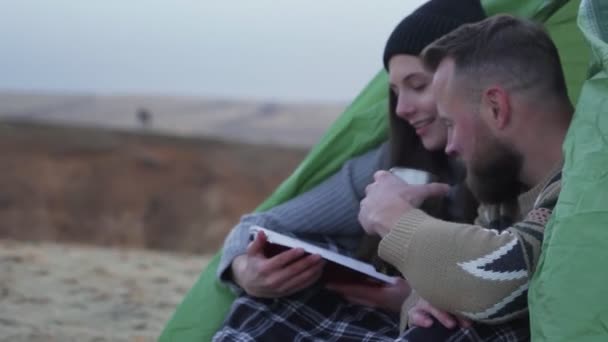 Vue latérale de yong homme et femme assis près de la tente et la voiture, boit un thé chaud et lire le livre dans la montagne — Video