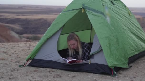 Молодая женщина лежит в палатке и читает книгу на горе — стоковое видео