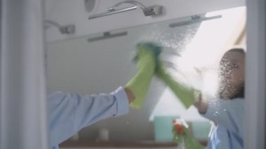 Banyodaki genç kadın aynayı kristal spreyle yıka ve yeşil bir bezle temizle.