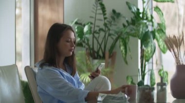 Mutfakta masada oturan ve akıllı telefon kullanan genç bir kadın. Dinamik kamera hareket ediyor.