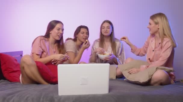 Cuatro jóvenes guapas felices en pijama se sientan en la cama, comen palomitas de maíz y ven una película en la despedida de soltera — Vídeo de stock