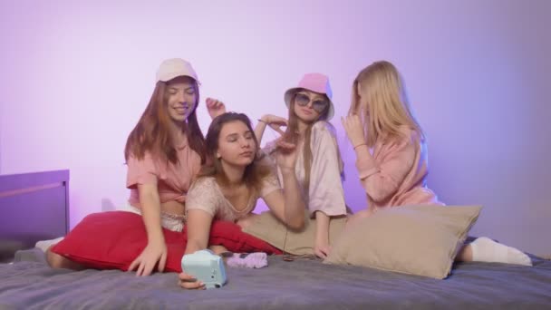 Четыре счастливые молодые женщины в пижаме сидят на кровати и берут серебро на винтажную камеру в солнечных очках и шляпах на девичнике — стоковое видео
