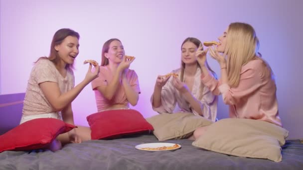 Fyra glada söta unga kvinnor i pyjamas sitter på sängen, äter pizza och tar bilder på möhippa — Stockvideo