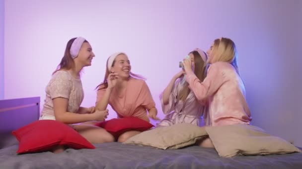 Cuatro hermosas mujeres jóvenes felices en pijama se sienta en la cama, se pone diademas y toma fotos en la despedida de soltera — Vídeos de Stock