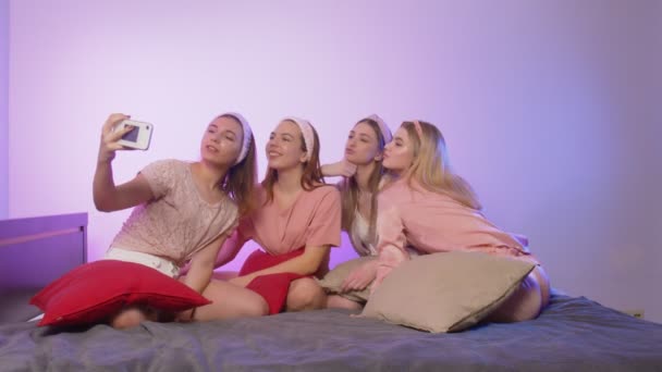 Cztery szczęśliwe, ładne, młode kobiety w piżamie, opaski i opaski na głowie siedzą na łóżku i robią zdjęcia przez telefon na wieczorze panieńskim. — Wideo stockowe