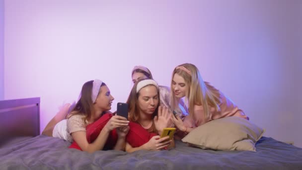 Four happy pretty young women in pajamas, Eye Collagen Patches and headbands on head sits on bed and uses phones at bachelorette party — Stock Video