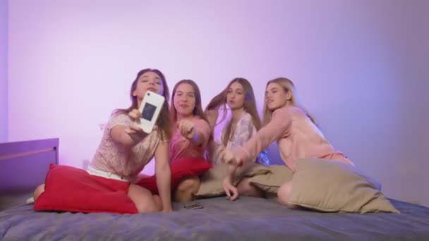 Four happy pretty young women in pajamas sits on bed, sings and records video on their phones at bachelorette party