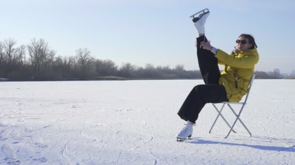 Mladá žena ve žluté bundě sedí na židli na zamrzlém jezeře a dává na bílé krasobruslení venku — Stock video