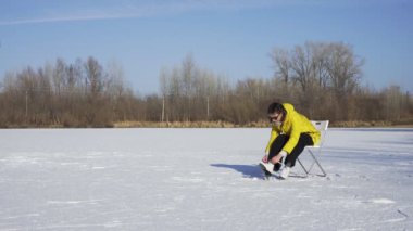 Güneş gözlüklü ve sarı ceketli genç bir kadın donmuş gölde sandalyede oturur ve açık havada beyaz bir buz pateni giyer.