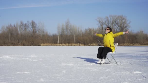 Młoda kobieta w okularach przeciwsłonecznych, żółtej kurtce i białej łyżwach figurowych siedzi na krześle na zamarzniętym jeziorze i pozuje na zewnątrz kamery — Wideo stockowe
