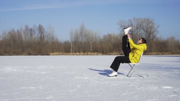 Mladá žena v slunečních brýlích a žluté bundě sedí na židli na zamrzlém jezeře a dává na bílé krasobruslení venku — Stock video