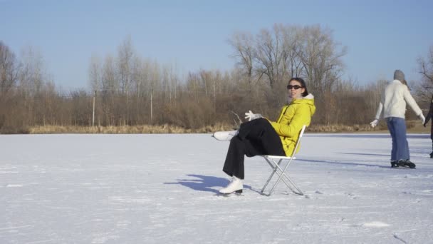 Mladá žena v slunečních brýlích, žluté bundě a bílých krasobruslích sedí na židli na zamrzlém jezeře a obléká si rukavice, zatímco její přátelé bruslí na pozadí — Stock video