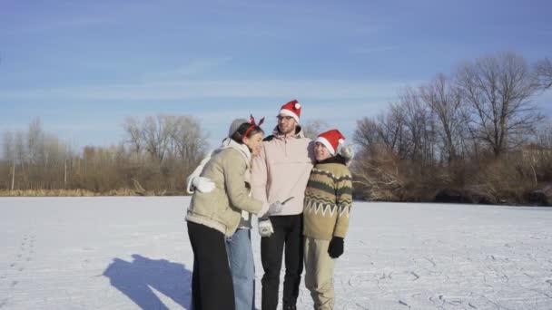 크리스마스 산타 모자를 쓴 네 명의 친구들은 숲 속에 있는 얼어붙은 큰 호수에서 빙판을 타고 난 뒤 휴대폰으로 셀카를 꺼내려 한다. — 비디오