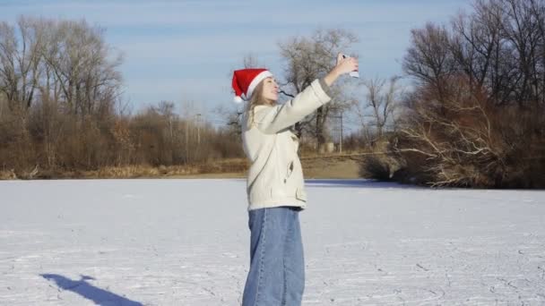 Young woman in christmas santa hat rides ice skating and takes selfie on phone on frozen lake in forest — Stock Video