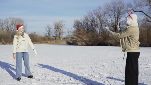 Mladá žena v vánoční santa klobouk vezme fotografii své přítelkyně na telefonu na zamrzlé jezero v lese — Stock video