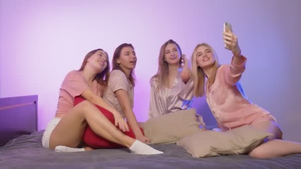 Vier gelukkige mooie jonge vrouwen in pyjama zit op bed en neemt selfies op de telefoon op vrijgezellenfeest — Stockvideo