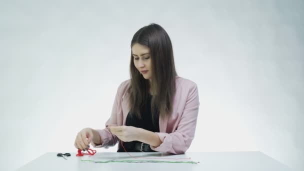 Vonzó fiatal nő sző egy karkötőt az asztalnál egy fehér stúdióban — Stock videók