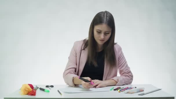 Mujer joven atractiva utiliza un marcador rosa para dibujar algo en un papel blanco en la mesa en un estudio blanco — Vídeos de Stock