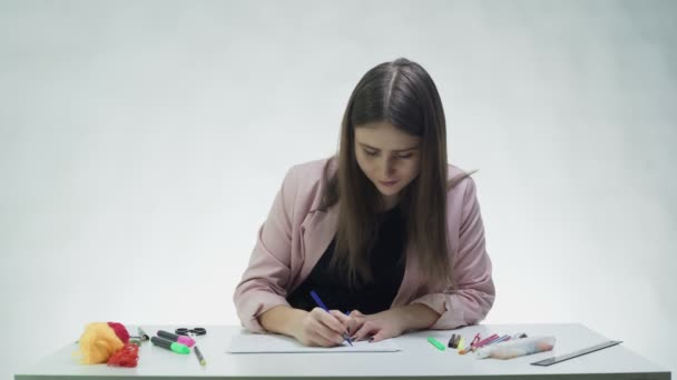 Mujer joven atractiva utiliza marcadores para dibujar en un papel blanco en la mesa en un estudio blanco — Vídeo de stock
