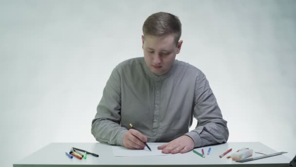 Jongeman tekent een meisje gezicht op papier aan tafel in een witte studio — Stockvideo