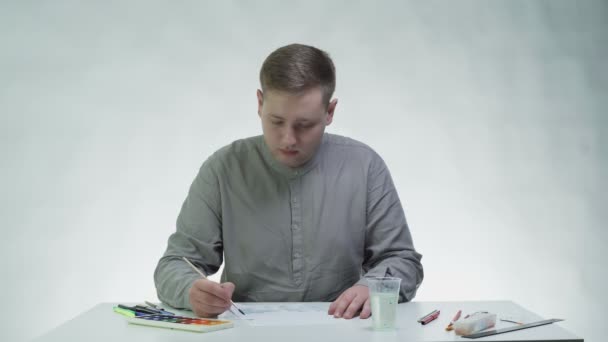 Young man draws with watercolors on a white paper at the table in a white studio — Stock Video