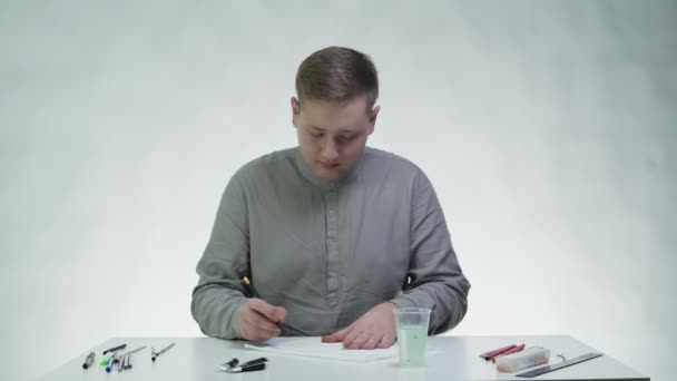 Jongeman tekent met een rode acrylverf op een witte doek aan tafel in een witte studio — Stockvideo