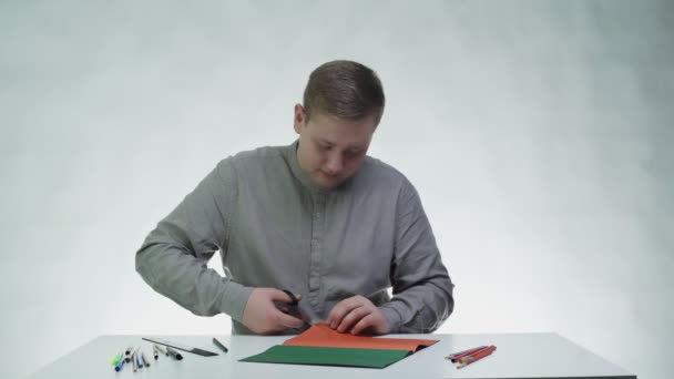 Młody człowiek używa nożyczek do cięcia pomarańczowego papieru przy stole w białym studiu. — Wideo stockowe