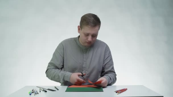 Młody człowiek używa nożyczek do wycinania czegoś z pomarańczowego papieru przy stole w białym studiu. — Wideo stockowe