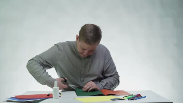 Jongeman gebruikt een schaar om een groene kleur papier te knippen aan de tafel in een witte studio — Stockvideo