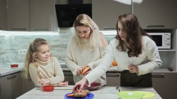 Die junge Mutter und ihre beiden Töchter kochen in der Küche Salat. Frau und Tochter putzen Karotten und Kartoffeln. — Stockvideo