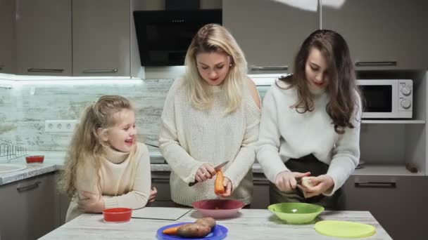 Die junge Mutter und ihre beiden Töchter kochen in der Küche Salat. Frau und Tochter putzen Karotten und Kartoffeln und reden — Stockvideo