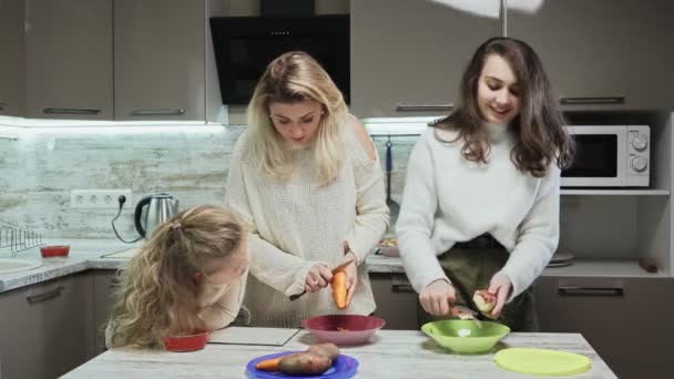 Die junge Mutter und ihre beiden Töchter kochen in der Küche Salat. Frau und Tochter putzen Karotten und Kartoffeln und reden — Stockvideo