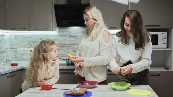 Die junge, glückliche Mutter und ihre beiden Töchter kochen in der Küche Salat. Kleine Tochter wäscht Möhren unter Wasser und älteste Tochter putzt Kartoffeln — Stockvideo