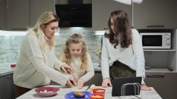 Die junge Mutter und ihre beiden Töchter kochen in der Küche Salat. Mutter bringt ihrer kleinen Tochter bei, wie man Karotten schneidet — Stockvideo