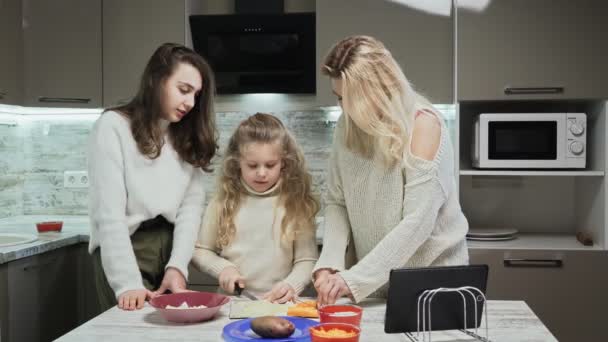 Die junge Mutter und ihre beiden Töchter kochen in der Küche Salat. Mutter ihrer kleinen Tochter schneidet Gemüse, während älteste Tochter mit ihnen spricht — Stockvideo