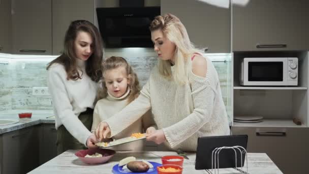 Die junge Mutter und ihre beiden Töchter kochen in der Küche Salat. Mutter und kleine Tochter legen geschnittene Kartoffeln in Teller — Stockvideo