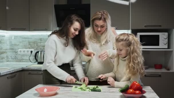 Ung mor och två döttrar lagar sallad i köket. Mor och döttrar skär gurkor — Stockvideo