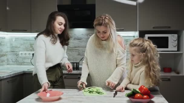 Die junge Mutter und ihre beiden Töchter kochen in der Küche Salat. Mutter und ihre Töchter legen geschnittene Gurken in Teller — Stockvideo