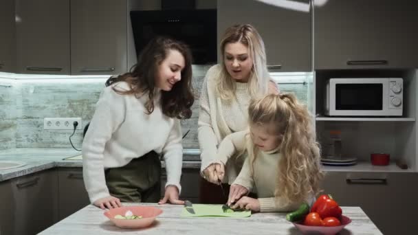 Νεαρή μητέρα και δύο κόρες της μαγειρεύουν σαλάτα στην κουζίνα. Η μητέρα μαθαίνει στην κορούλα της να κόβει αγγούρια. — Αρχείο Βίντεο