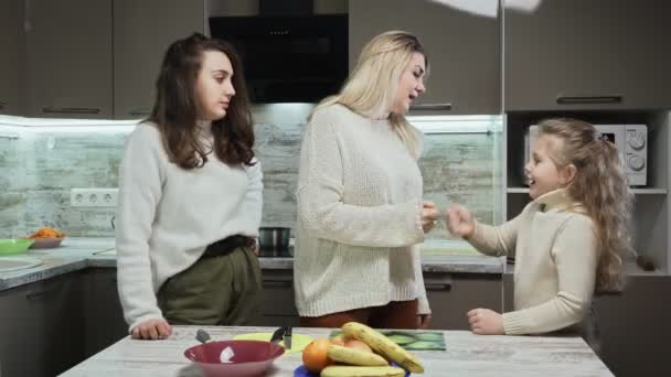 Junge Mutter mit zwei Töchtern kocht Obstsalat in Küche. — Stockvideo