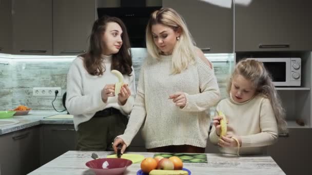Ibu muda dan dua putrinya memasak salad buah di dapur. Sisters peel pisang dan mempersiapkan mereka untuk memotong dalam salat buah — Stok Video