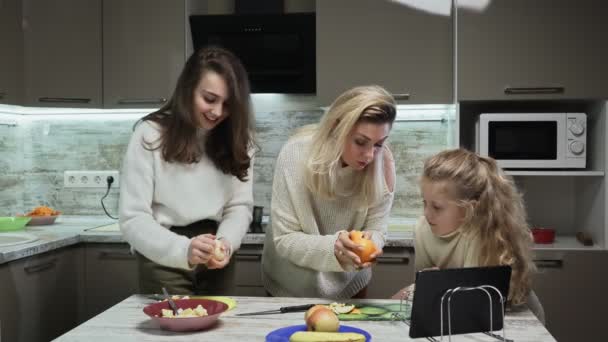 Νεαρή μητέρα και δύο κόρες της μαγειρεύουν φρουτοσαλάτα στην κουζίνα. Οι μητέρες δείχνουν πώς να ξεφλουδίζουν το πορτοκάλι στην κορούλα της. — Αρχείο Βίντεο