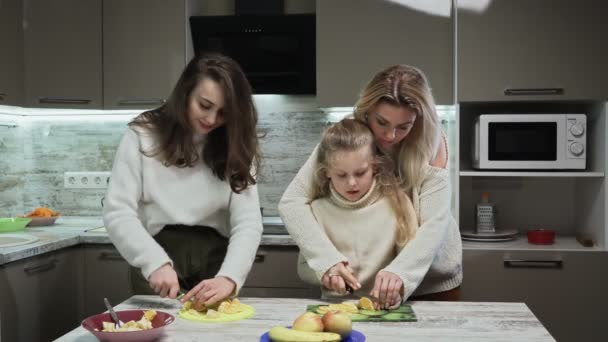 Νεαρή μητέρα και δύο κόρες της μαγειρεύουν φρουτοσαλάτα στην κουζίνα. Η μητέρα μαθαίνει στην κορούλα της να κόβει πορτοκάλι. — Αρχείο Βίντεο