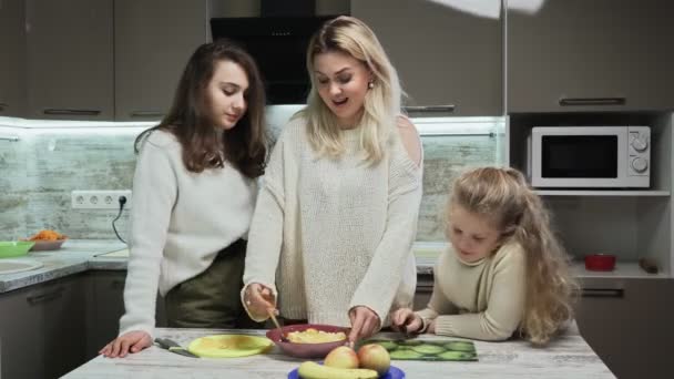 Die junge Mutter und ihre beiden Töchter kochen in der Küche Obstsalat. Mutter mischt Obstsalat mit Bananen und Orangen — Stockvideo