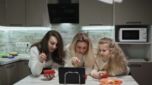 Η νεαρή μητέρα και οι δύο κόρες της τρώνε σαλάτα και βλέπουν σε μια ταμπλέτα στην κουζίνα.. — Αρχείο Βίντεο