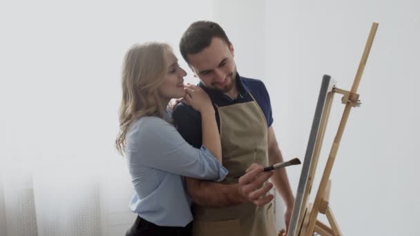 Joven artista masculino con pinturas de delantal en lienzo dibujo con su novia en el estudio de arte. — Vídeo de stock