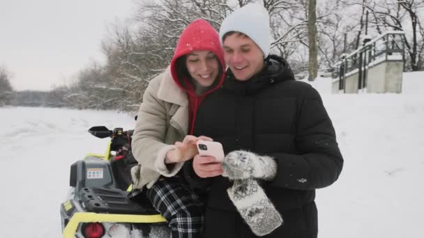 Jong stel poseren met ATV Quad fiets in het winterbos. Jongeman en vrouw kijkt naar de telefoon — Stockvideo